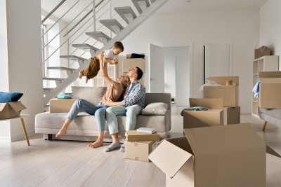Happy young family couple playing with small toddler kid son together in new home. Parents and child boy having fun in new house living room with boxes. Moving day, mortgage, relocation concept