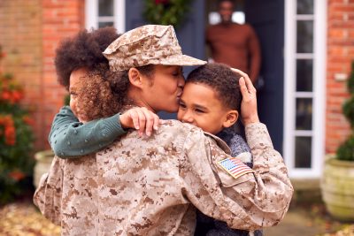 American Female Soldier In Uniform Returning Home To Family On Hugging Children Outside House